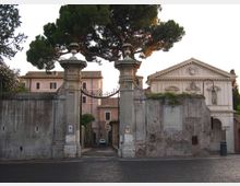 Ingresso monumentale con due pilastri in pietra e cancello in ferro battuto inseriti in un alto muro, con vialetto che conduce a edifici storici interni. Sullo sfondo si vedono grandi pini e una facciata in stile classico con frontone triangolare.