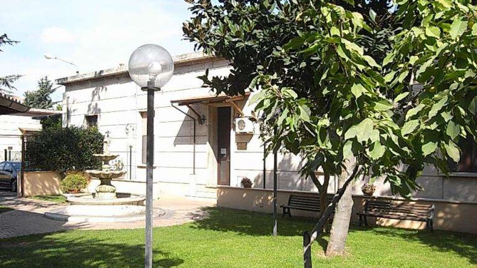 Cortile con prato verde e alberi, con lampione sferico al centro e panchine lungo il muro. Sullo sfondo si vede un edificio chiaro a un piano con ingresso e, a sinistra, una piccola fontana decorativa.