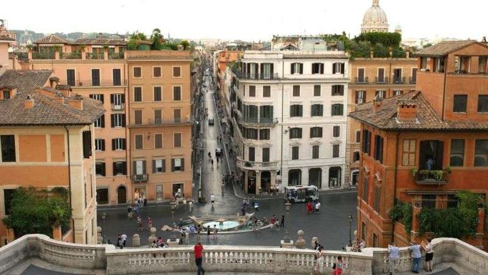 Veduta dall’alto di una piazza urbana con una fontana centrale e una lunga strada rettilinea che si estende tra palazzi storici dai toni ocra e bianco. In primo piano si vede una balaustra in pietra e, sullo sfondo, i tetti della città con una grande cupola visibile in lontananza.