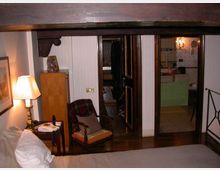 Interno di una camera da letto con soffitto a travi e arredi in legno, comprensivo di letto in primo piano, comodino con lampada e poltrona. Sullo sfondo si vede un corridoio con porte in legno che conduce a un bagno con piastrelle chiare e mobile lavabo verde.