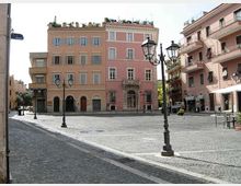 Piazza urbana pavimentata in sampietrini con lampioni in ferro battuto, circondata da palazzi a più piani color rosa con persiane e balconi. Al centro si nota un edificio con ingresso ad arco e finestre regolari, affiancato da altri edifici residenziali con negozi al piano terra.