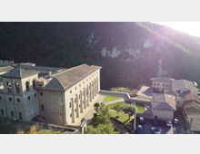 Complesso di edifici in pietra con tetti in tegole, dominato da un grande edificio rettangolare con alte finestre ad arco, affacciato su un cortile e aree verdi. Sullo sfondo si vede un pendio montuoso boscoso, con strada e piccoli edifici adiacenti.