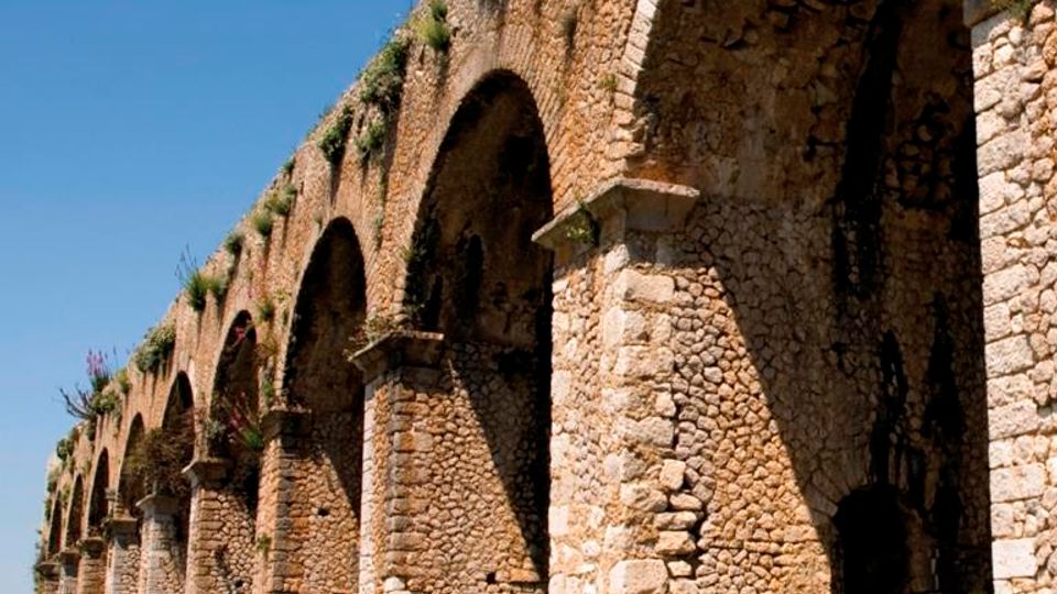 Antico acquedotto in pietra con una serie di grandi archi e pilastri, visto in prospettiva lungo un prato. La struttura in muratura grezza presenta vegetazione che spunta dalla sommità sotto un cielo azzurro.
