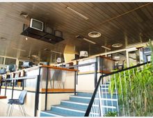 Interno di un ufficio open space su due livelli con scale e corrimano in metallo, postazioni con computer e grandi vetrate lungo il perimetro. Soffitto a pannelli con luci fluorescenti, diffusori d’aria e vecchi monitor sospesi, con piante ornamentali vicino alla scala.