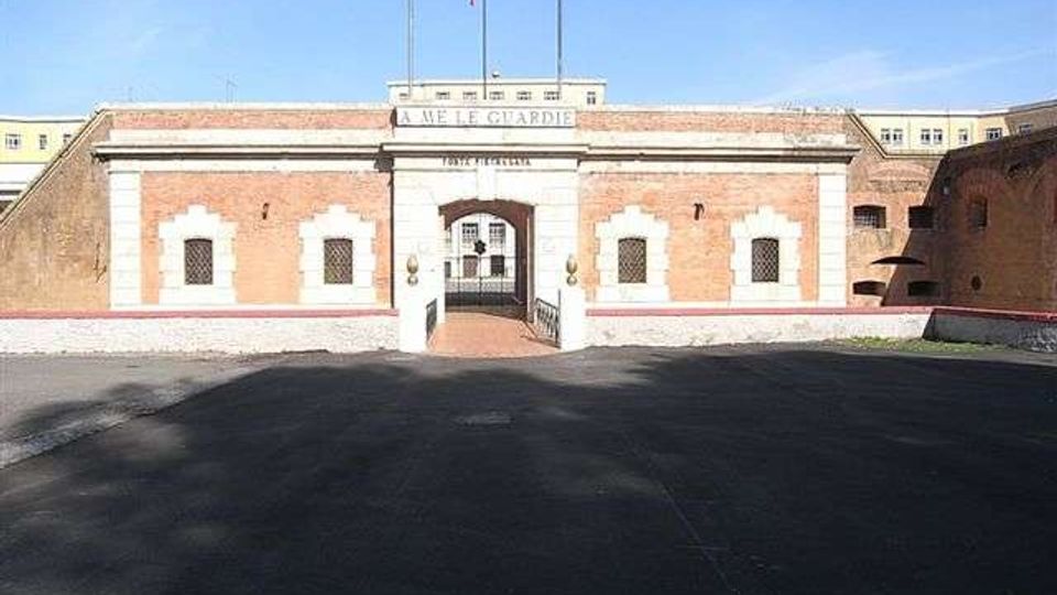 Facciata simmetrica in mattoni rossi con cornici in pietra bianca e ingresso ad arco centrale con cancello, sormontato da un’insegna e da due aste portabandiera. Davanti si estende un ampio piazzale asfaltato, con mura laterali che incorniciano l’accesso.