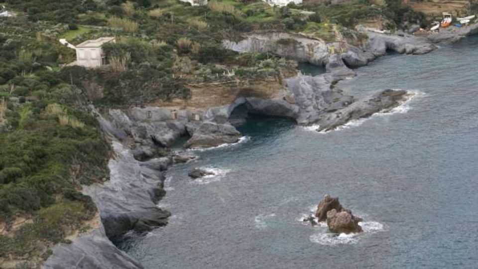 Veduta dall’alto di una costa rocciosa con falesie grigie erose e una grande grotta marina affacciata su una piccola insenatura. Nell’entroterra si vedono vegetazione mediterranea, campi terrazzati e alcune case bianche sparse vicino alla scogliera.