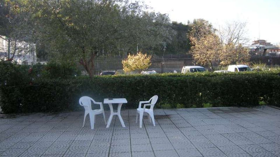 Terrazza pavimentata con piastrelle a motivi geometrici, arredata con un piccolo tavolo e due sedie di plastica bianche. Sullo sfondo ci sono una siepe, alberi e una strada con auto, con una collina boscosa e edifici lontani.