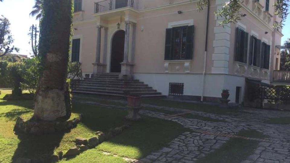 Villa su due piani con facciata chiara, finestre con persiane scure e ingresso ad arco con colonne, raggiunto da una breve scalinata. Davanti si vede un giardino con prato, palme e un vialetto carrabile in pietra che conduce all’edificio.