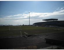 Campo da calcio all’aperto con pista perimetrale e piccole porte, illuminato da un alto palo faro. Sul lato destro si vede una tribuna coperta e bassa, con il cielo aperto sullo sfondo.
