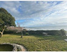 Giardino costiero con prato, siepi basse e alcuni alberi, affacciato su una spiaggia e sul mare con piccole onde. In primo piano si vede una panchina in pietra e un piccolo stagno delimitato da pietre, sotto un cielo parzialmente nuvoloso.
