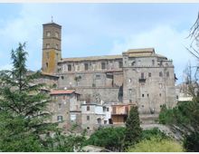 Complesso storico in pietra su un’altura, con alta torre campanaria quadrata e corpi di edificio fortificati con piccole finestre e tetti in coppi. In primo piano vegetazione e alberi, sullo sfondo cielo chiaro.