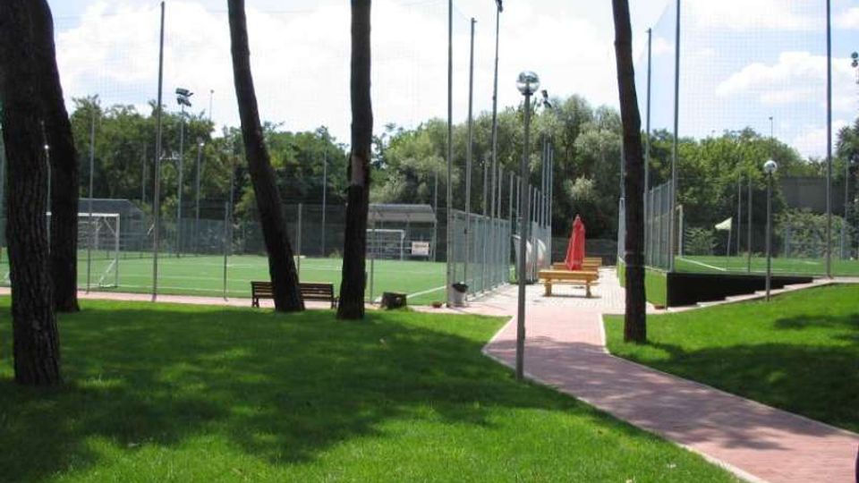 Parco sportivo con campi da calcio in erba sintetica recintati da alte reti e pali di illuminazione. In primo piano un prato con alberi e una passerella pavimentata con panchine e un ombrellone chiuso, circondati da vegetazione.