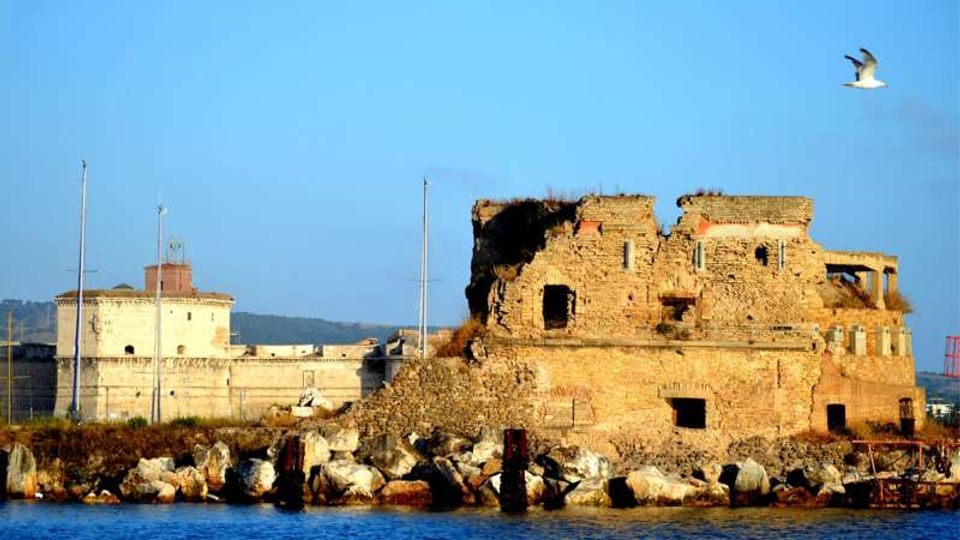 Rovine di un edificio in muratura in riva al mare, con muri in pietra parzialmente crollati e aperture ad arco, circondate da massi frangiflutti. Sullo sfondo si vede una struttura fortificata chiara con torrette e aste per bandiere, sotto un cielo azzurro.
