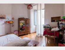 Interno di una camera da letto con pareti rivestite di carta da parati chiara, pavimento in parquet e grande finestra con tende bianche e persiane aperte. Arredi in legno includono un letto, un armadio e un tavolo con sedie, con mensole e oggetti domestici lungo le pareti.