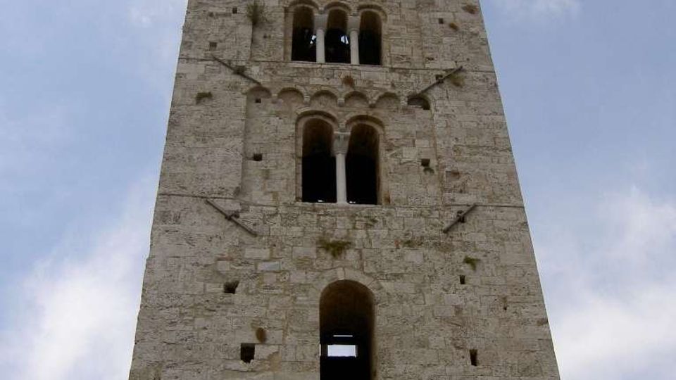 Torre medievale in pietra a pianta rettangolare, ripresa dal basso, con più livelli di finestre ad arco e aperture geminate sovrapposte. La facciata presenta mensole e tiranti metallici visibili, con cielo parzialmente nuvoloso sullo sfondo.