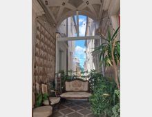 Piccolo atrio coperto con volta decorata e pareti a motivi geometrici, arredato con divanetto e sedie in stile classico tra molte piante. In fondo una grande vetrata ad arco lascia vedere una stretta via tra edifici in pietra sotto il cielo.