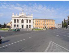 Veduta di una grande piazza urbana con ampia carreggiata e spartitraffico, dominata da una facciata monumentale in stile neoclassico con colonne, arcate e statue sul coronamento. Sullo sfondo si vedono edifici storici allineati lungo la strada, con cielo parzialmente nuvoloso.