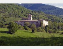 Edificio storico in pietra con pianta rettangolare e torre campanaria centrale, situato su un pendio tra prati verdi e alberi. Sullo sfondo si vedono colline e montagne boscose che circondano la struttura.