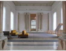 Ampia sala interna con alte colonne e soffitto decorato, pavimento in pietra e grandi finestre che illuminano lo spazio. Alcune sedute moderne e sedie sono disposte lungo le pareti, con un’apertura centrale che conduce a un’altra stanza con vetrata.