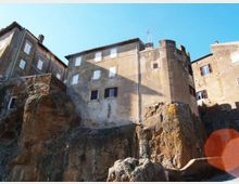 Case in pietra e intonaco di un borgo storico costruite sopra alte rocce, con finestre e una torre cilindrica. In basso si vedono pareti rocciose e un piccolo tratto di strada o passaggio, sotto un cielo sereno.