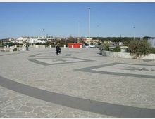 Ampia piazza pavimentata in pietra con grandi fasce geometriche scure e motivi decorativi incisi sul suolo, illuminata da alti lampioni. Sullo sfondo si vedono aiuole con vegetazione bassa, parcheggi e edifici bassi sotto un cielo sereno.