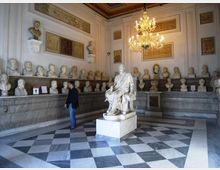 Sala museale neoclassica con pavimento a scacchi in marmo e pareti decorate con rilievi e pannelli, illuminata da un grande lampadario. Lungo il perimetro sono esposti numerosi busti in marmo su mensole, con una statua seduta su piedistallo al centro della stanza.