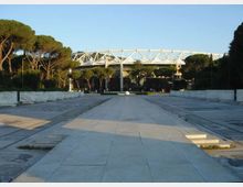 Ampio viale pedonale lastricato che conduce all’ingresso di uno stadio, con struttura superiore bianca a tralicci visibile sullo sfondo. Ai lati si trovano alberi alti e aree pavimentate con muretti bassi, sotto un cielo sereno.