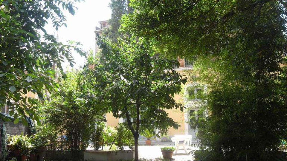 Cortile-giardino ombreggiato con ghiaia a terra e alberi e arbusti fitti; in primo piano si vede un tubo da giardino. Sullo sfondo compaiono una piccola area pavimentata con tavolino e vasi e la facciata di un edificio con finestre.