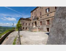 Cortile in cemento davanti a un grande edificio storico in pietra con finestre chiuse da persiane e un portale ad arco raggiungibile tramite una scalinata. Sul lato sinistro corre una strada con muretto e lampioni, affacciata su una valle collinare sotto cielo azzurro.