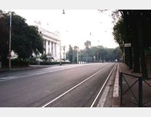 Ampio viale urbano con binari del tram al centro e marciapiede con ringhiera sul lato destro, fiancheggiato da alberi e lampioni. Sul lato sinistro si vede un grande edificio monumentale con facciata neoclassica a colonne e scalinata.