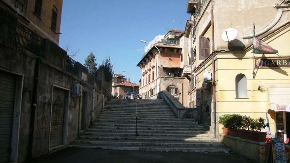 Scalinata in pietra in una strada urbana in salita, affiancata da edifici residenziali di più piani con facciate intonacate, balconi e persiane. Ai lati si vedono muri e saracinesche di negozi chiusi, lampioni stradali e un piccolo bar con insegna sulla destra.