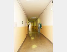 Lungo corridoio interno di un edificio, con pareti bianche e fascia inferiore gialla, pavimento lucido e illuminazione a neon sul soffitto. Ai lati si vedono porte e segnaletica di sicurezza, con un’uscita sul fondo del passaggio.
