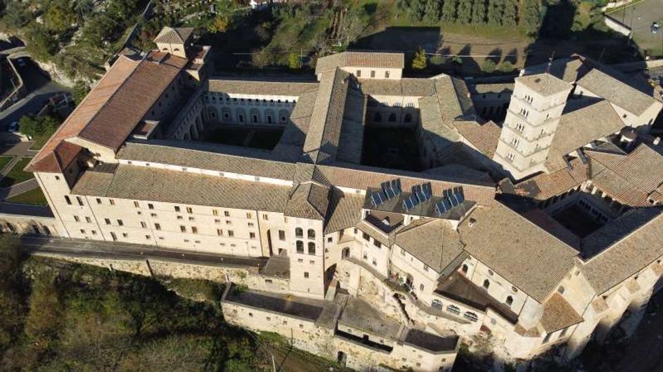 Veduta aerea di un grande complesso monastico in pietra con tetti in coppi, articolato attorno a più cortili interni e corridoi. Si notano un campanile quadrato e mura perimetrali, circondati da aree verdi e terreni agricoli.