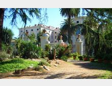 Viale sterrato in un giardino mediterraneo con palme, agavi e vegetazione fitta, che conduce a un cancello in ferro tra pilastri chiari con vasi ornamentali. Sullo sfondo si vede un grande edificio storico chiaro con più piani e finestre regolari.