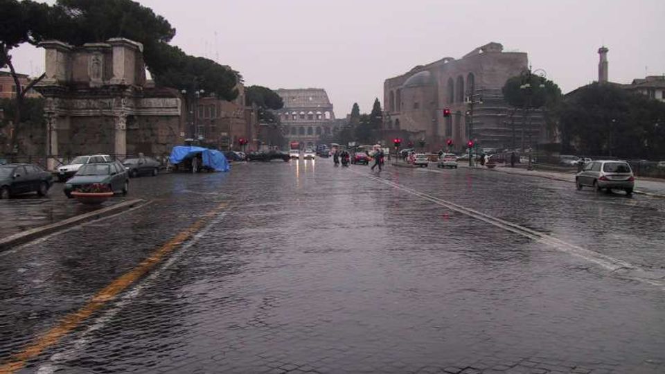 Ampio viale urbano in pavé bagnato dalla pioggia, con auto e semafori lungo la strada. Ai lati si vedono rovine e edifici storici con alberi, e in fondo un grande anfiteatro in pietra.