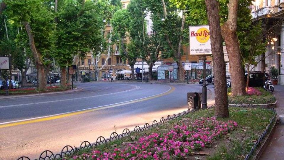 Viale cittadino alberato con strada curva e marciapiede, bordato da aiuole di fiori rosa e piccole recinzioni metalliche. Sullo sfondo si vedono edifici e fermate lungo la strada, con un’insegna “Hard Rock Cafe” sulla destra.