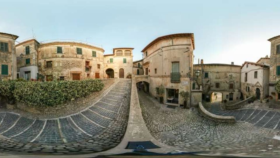 Piazza in un borgo storico con pavimentazione in pietra e gradinate curve che scendono verso un’area centrale. Intorno si vedono edifici antichi in muratura intonacata e pietra, con finestre e balconi, e passaggi ad arco tra le case.