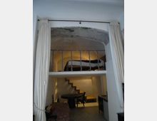 Interno di un piccolo appartamento con soppalco: in basso zona soggiorno con tavolo rotondo e scala a gradini che porta al livello superiore. Il soppalco ha una ringhiera metallica e un soffitto ad arco in pietra, con tende chiare ai lati dell’apertura.