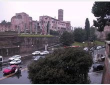 Veduta urbana con un complesso storico in mattoni e una torre sullo sfondo, affiancato da rovine con colonne e mura antiche. In primo piano si vede una strada bagnata dalla pioggia con auto in transito e aree verdi con alberi.