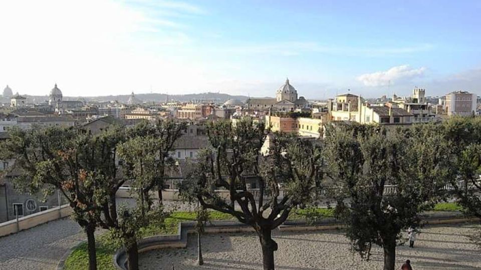 Veduta panoramica su una città con numerose cupole e campanili all’orizzonte, sotto un cielo limpido. In primo piano si vede un belvedere con alberi potati, vialetti curvi in pietra e una zona verde.