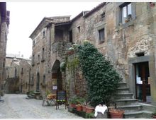 Stradina acciottolata in un borgo storico con edifici in pietra e intonaco, archi e balcone al piano superiore. Vasi con piante e un grande arbusto vicino a una scalinata esterna e a un ingresso con porta vetrata.