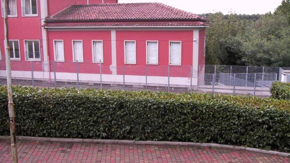 Edificio a due piani con facciata rosa e finestre rettangolari, affiancato da un corpo più basso con tetto in tegole. In primo piano ci sono una siepe e un vialetto in mattoni, con recinzione metallica e alberi sullo sfondo.