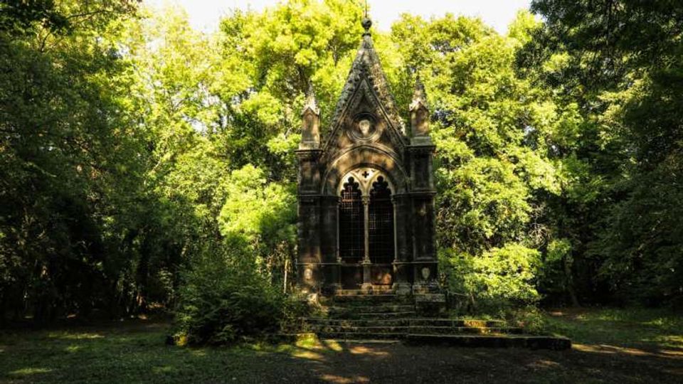 Piccola cappella in pietra in stile gotico, con frontone a punta, pinnacoli e grande finestra ad arco con grata, situata in una radura. Intorno si vede un fitto bosco di alberi e vegetazione che circonda l’edificio e l’area erbosa.