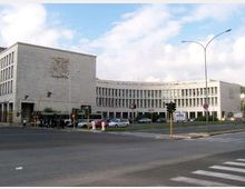 Grande edificio istituzionale moderno in pietra chiara, con un corpo principale rettangolare e un’ala curva a più piani con file regolari di finestre. In primo piano si vede un ampio incrocio urbano con semafori, strisce pedonali, auto parcheggiate e lampioni sotto un cielo parzialmente nuvoloso.