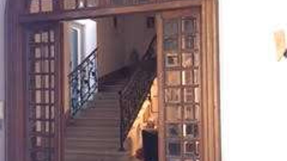 Grande portale ad arco in legno con ante a vetri che si apre su una scala interna in pietra con ringhiera in ferro battuto, in un ambiente dai muri chiari e pavimento piastrellato decorato.