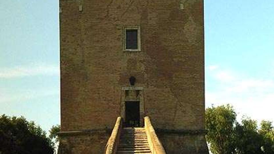 Torre in mattoni a pianta quadrata con sommità merlata e piccole finestre, raggiunta da una lunga scalinata centrale che conduce all’ingresso sopraelevato. La struttura è circondata da un prato verde con alberi ai lati sotto un cielo sereno.