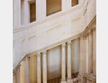 Interno di un edificio storico con ampia scalinata in pietra chiara che sale in diagonale, sostenuta da una fila di colonne. Pareti intonacate con riquadri decorativi e aperture superiori con ringhiere affacciano sul vano scala.