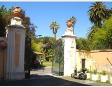 Ingresso di un parco o giardino con due alti pilastri in muratura chiara sormontati da vasi ornamentali e un cancello in ferro battuto aperto. All’interno si vedono vialetti e vegetazione con alte palme, mentre a destra corre un muro color ocra lungo la strada.
