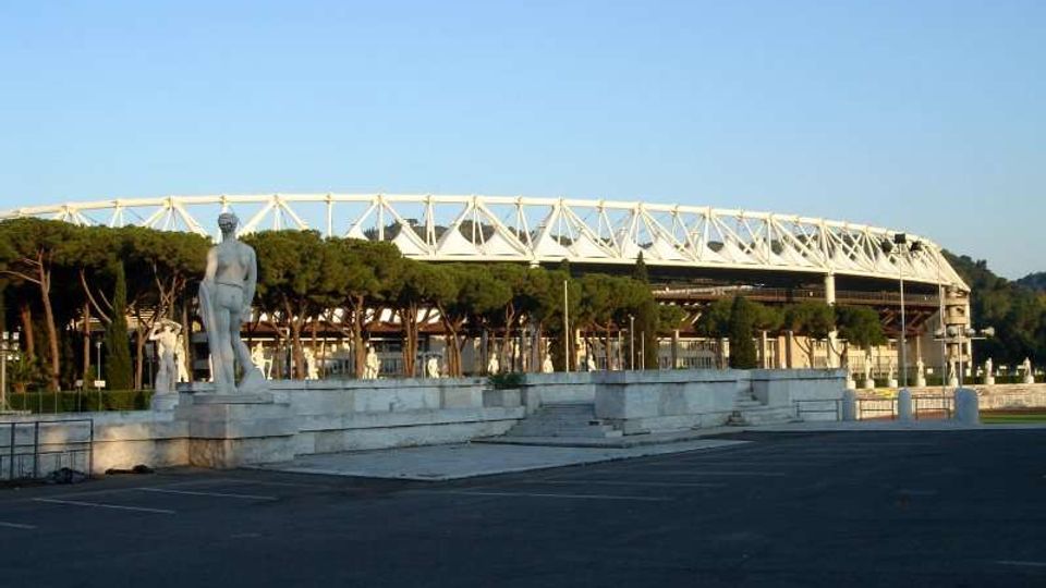 Veduta esterna di uno stadio con struttura ad anello e tralicci bianchi, sullo sfondo di un filare di pini. In primo piano si vede un’ampia piazza asfaltata con gradinate in pietra e una statua su piedistallo.