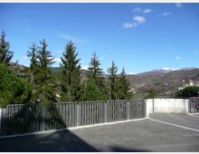 Terrazza o area pavimentata con ringhiera metallica lungo il bordo, affacciata su una valle con alberi sempreverdi e colline montuose. Sullo sfondo si vedono rilievi con chiazze di neve e un cielo azzurro con poche nuvole.